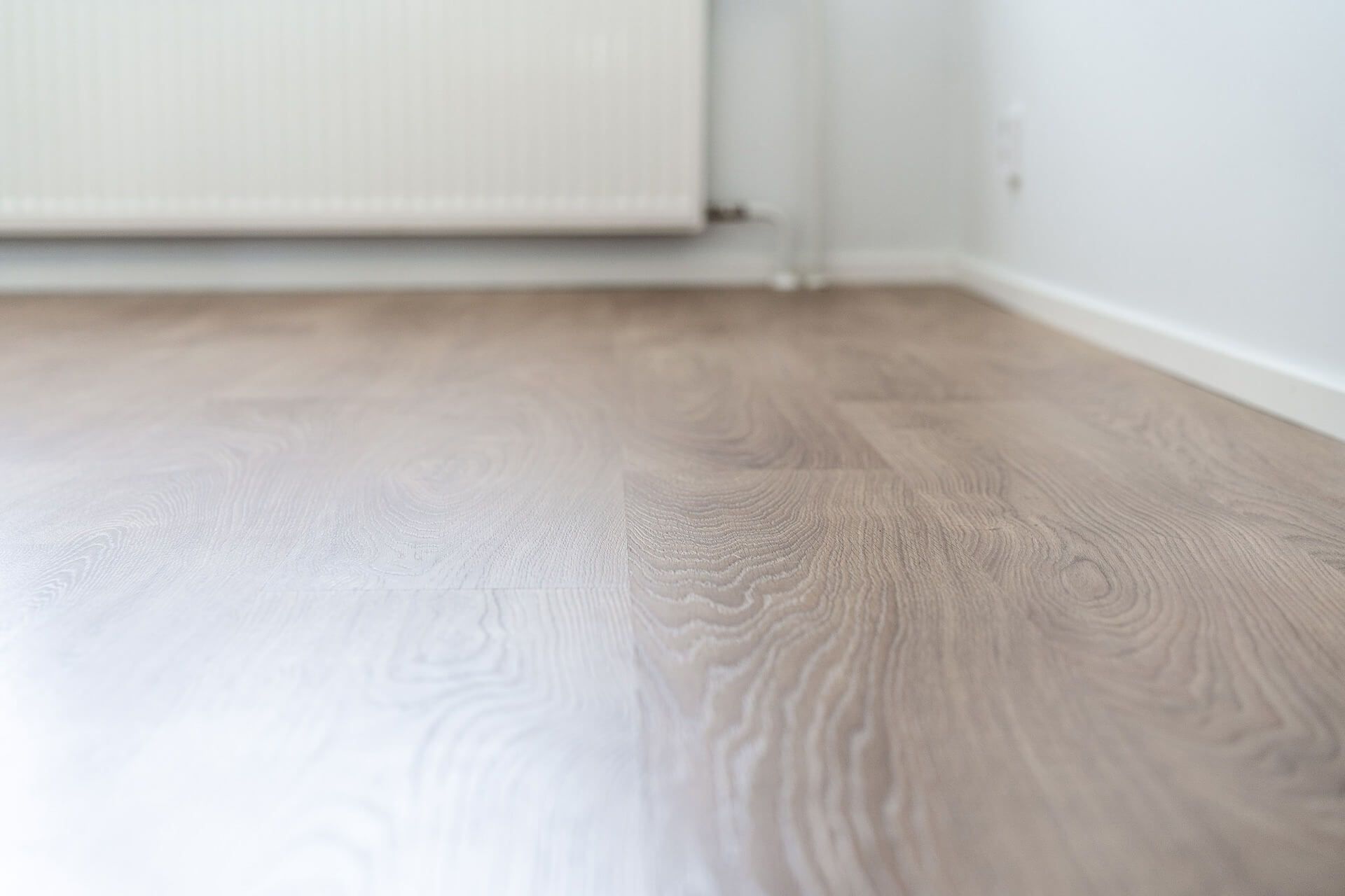 Vaaleanruskea laminaattilattia lähikuvassa, taustalla valkoinen patteri ja seinä.