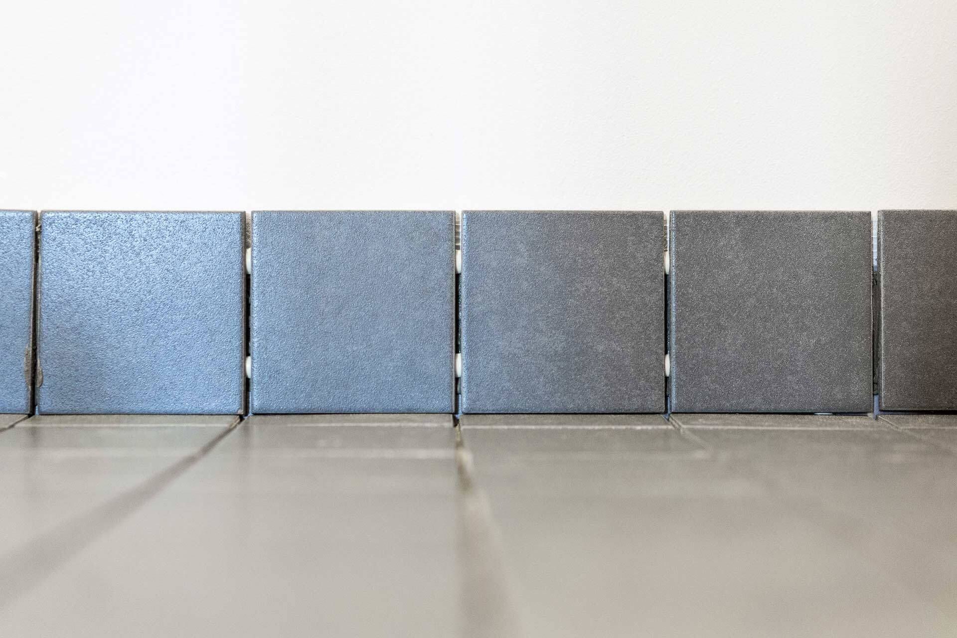 Rivi toisiinsa liitettyjä harmaita, kuvioituja neliönmuotoisia laattoja kiiltävällä lattialla valkoista seinää vasten.