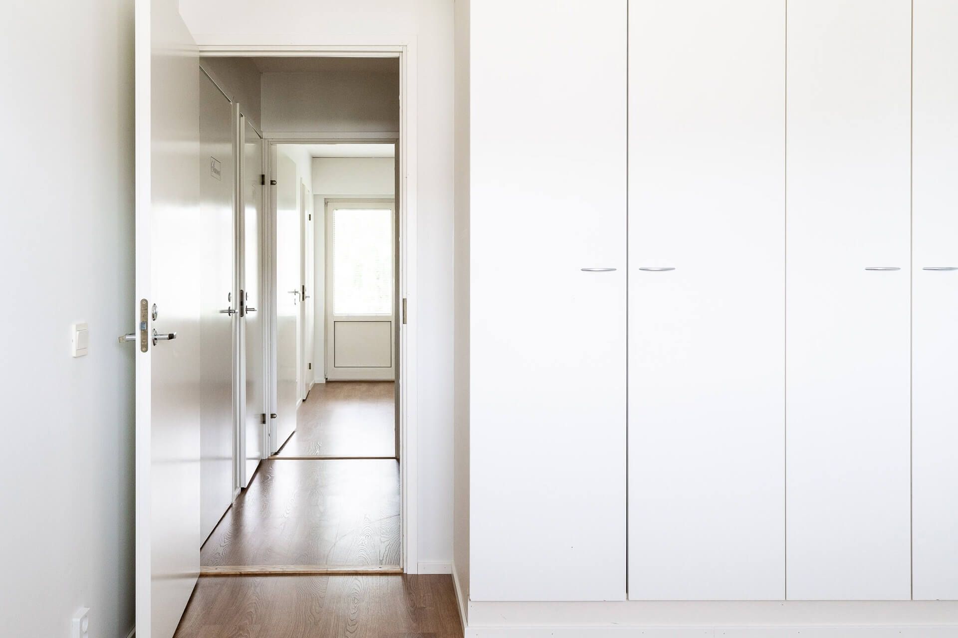 Käytävä, jossa on avoin oviaukko, valkoiset seinät ja puulattia. Kirkas luonnonvalo käytävän päässä.