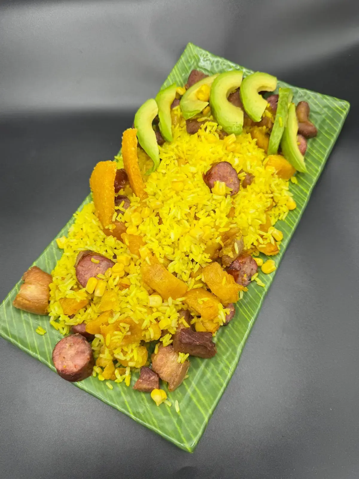 Un plato rectangular verde con arroz amarillo mezclado con salchicha, calabaza y frijoles, coronado con rodajas de aguacate.