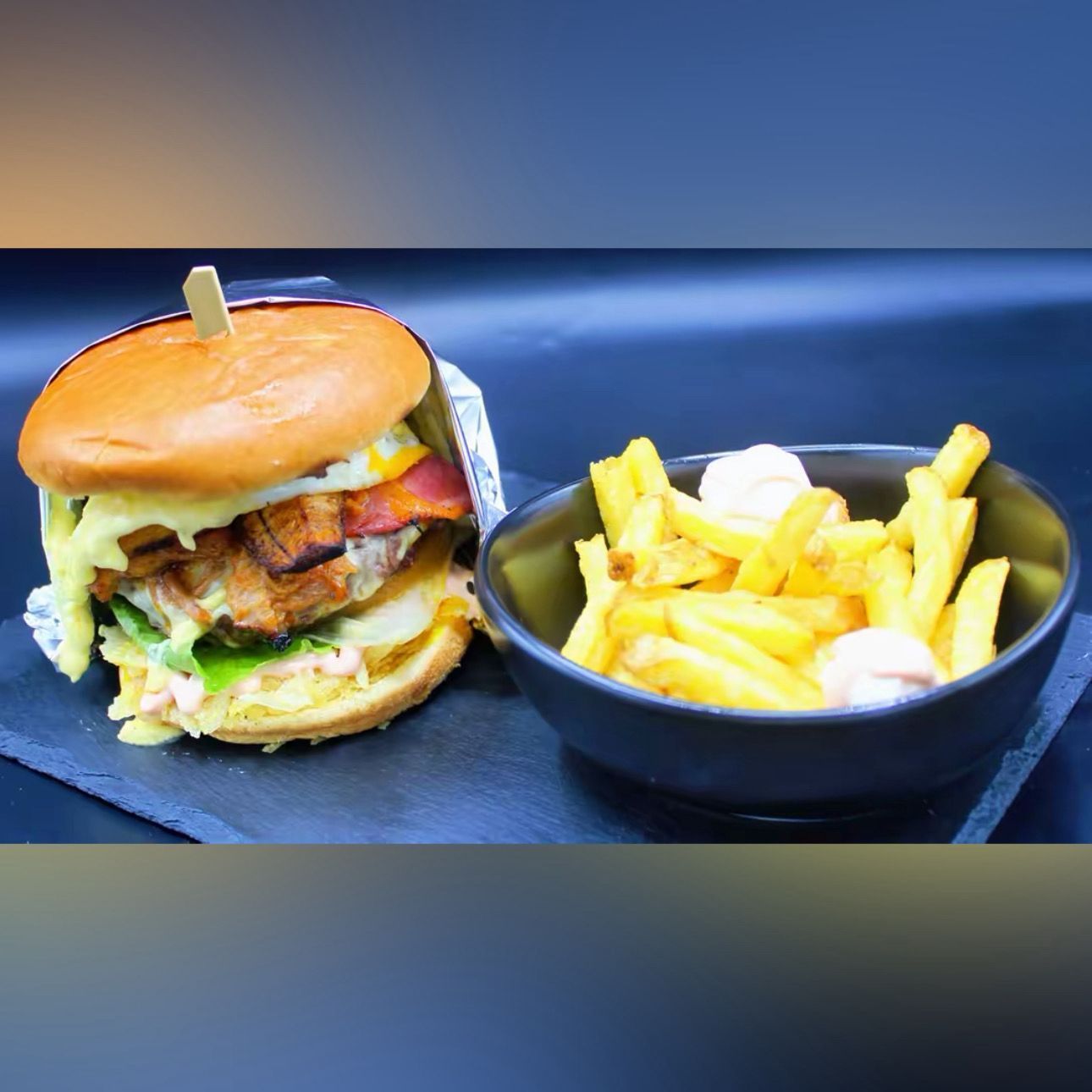 Una hamburguesa con queso y beicon, acompañada de patatas fritas con un poco de salsa, servida en un bol negro
