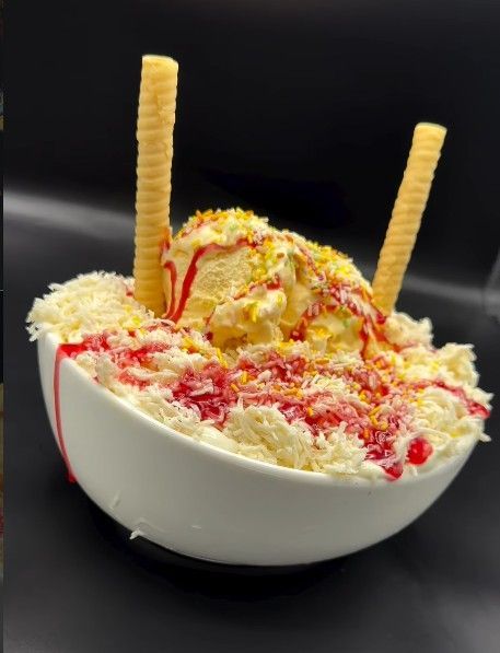 Un tazón de hielo raspado cubierto con helado de vainilla, jarabe rojo, coco rallado y dos barquillos, sobre un fondo negro.