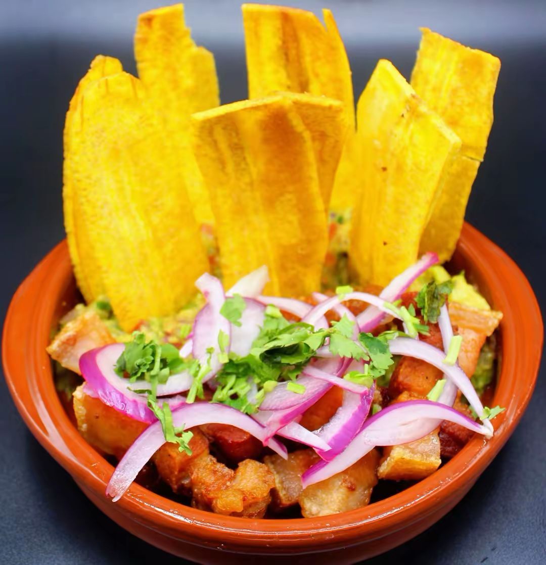 Un cuenco de barro con chicharrón y guacamole, cubierto con cebolla roja en rodajas y cilantro, servido con plátanos fritos.