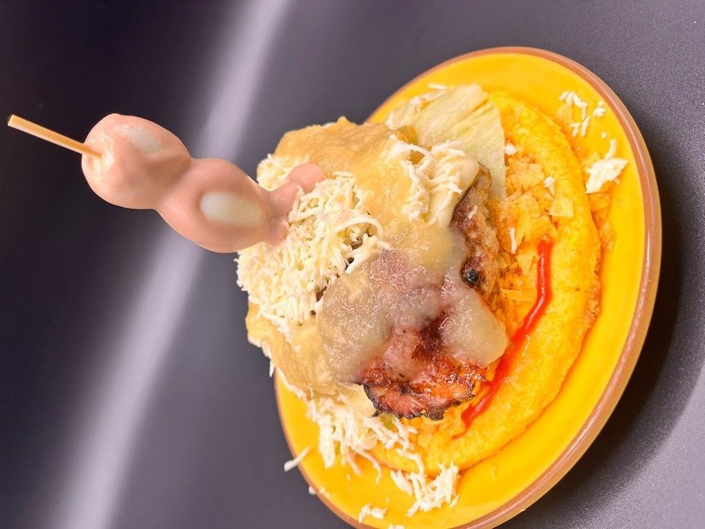 En un plato amarillo se sirve una hamburguesa cubierta con queso blanco rallado y salsa