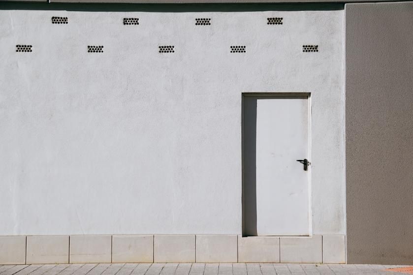 Pared blanca con puerta blanca; rejillas de ventilación rectangulares en la parte superior. Acera beige.