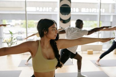 Mujer en clase de yoga, con los brazos extendidos en postura del guerrero, mirando a un lado. Otras personas al fondo.