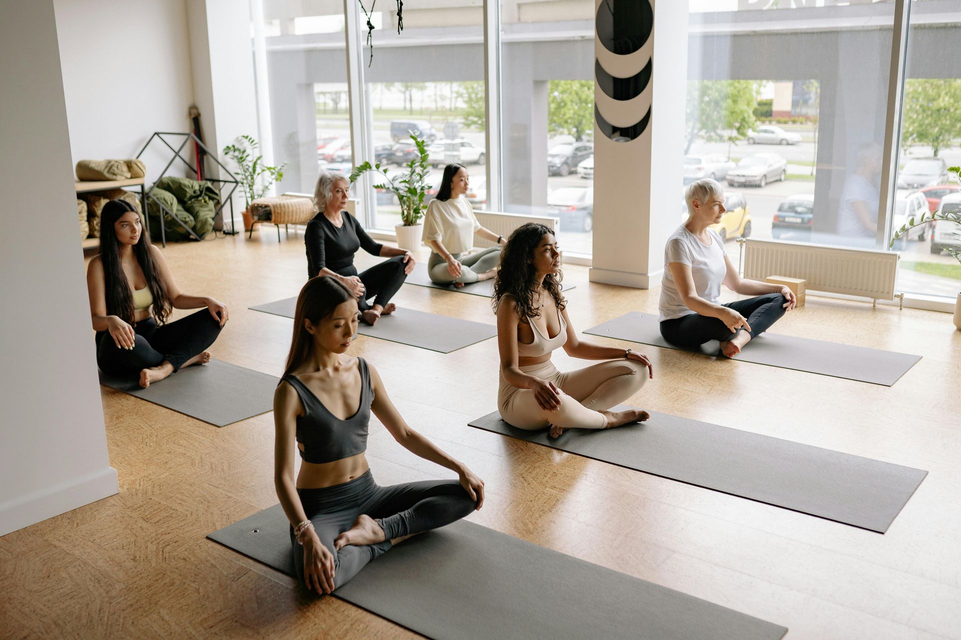 Mujeres en clase de yoga, sentadas con las piernas cruzadas sobre colchonetas, practicando meditación en un estudio luminoso.