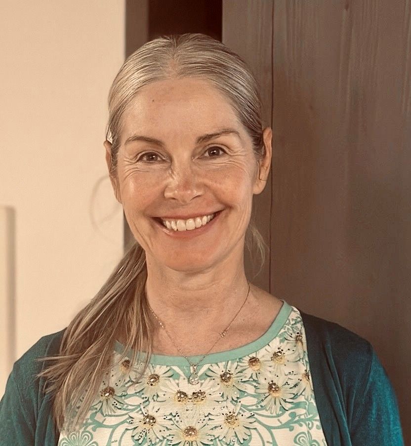 Frau mit ergrauendem Haar, lächelnd, trägt eine türkisfarbene Strickjacke über einem gemusterten Oberteil.