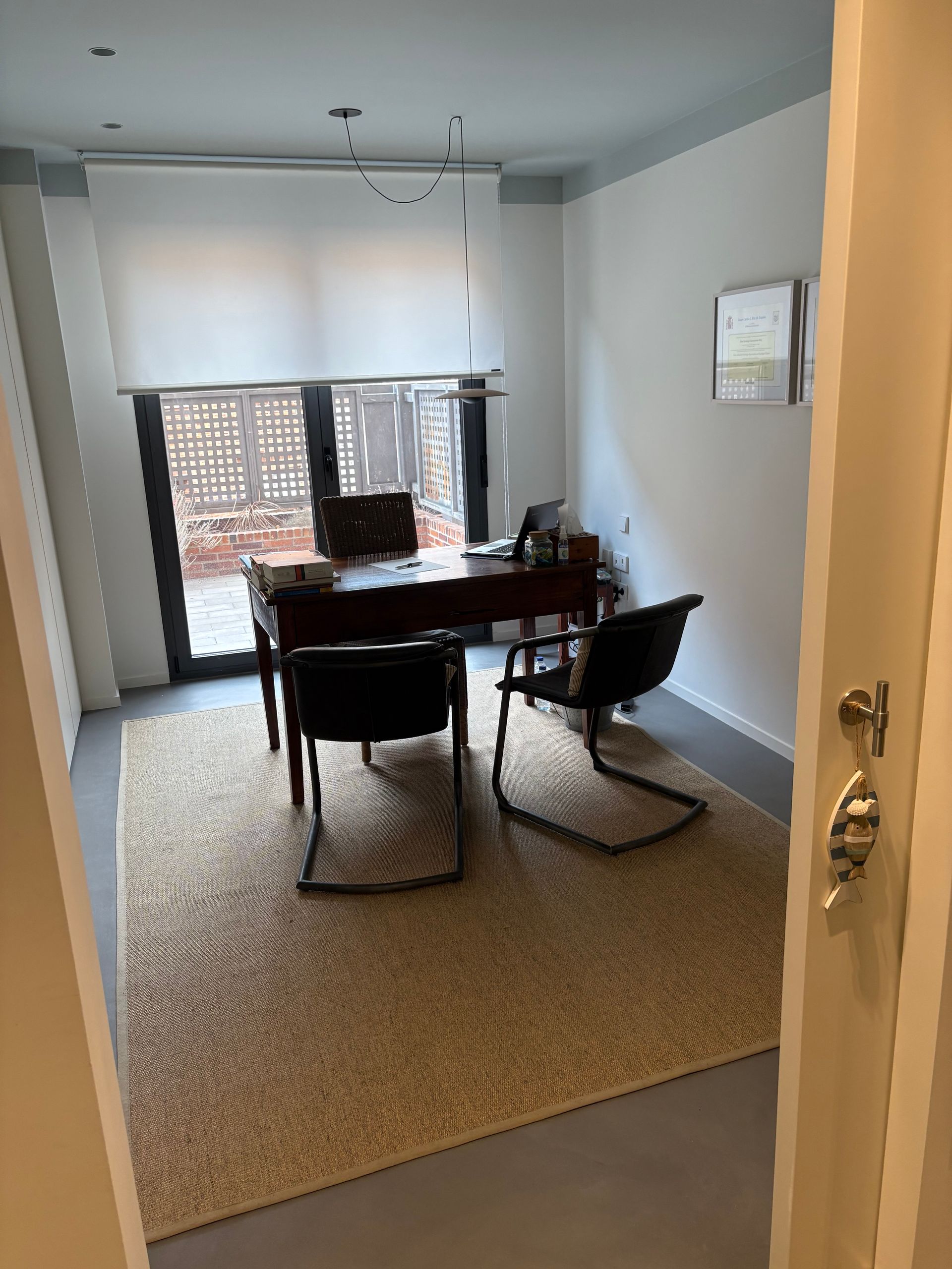 Oficina con escritorio, sillas, alfombra y acceso al exterior. Paredes blancas, techo azul y persianas que filtran la luz.