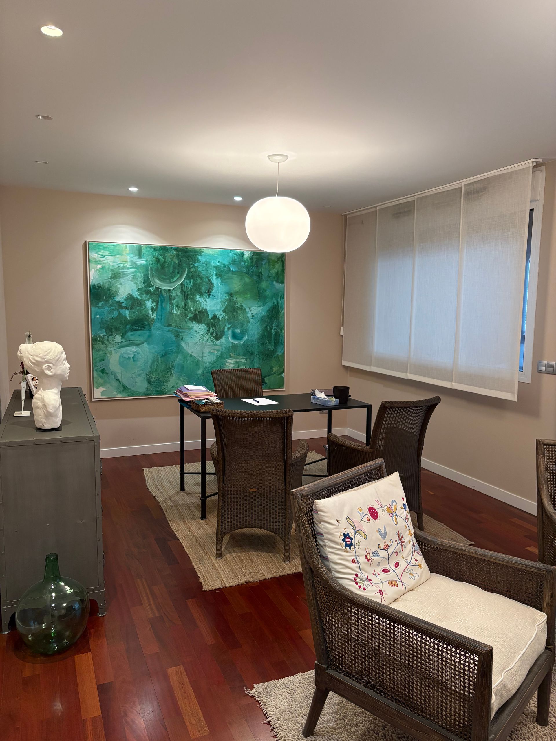 Habitación interior con mesa, sillas, obras de arte y lámpara colgante. Suelo de madera y paredes en tonos neutros.