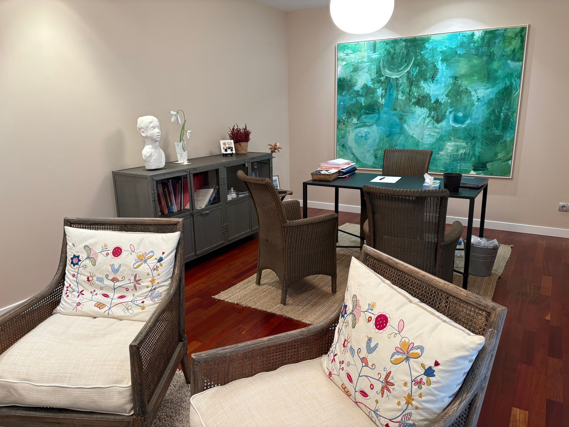 Interior de oficina con escritorio, sillas, obras de arte y armario. Paredes color beige, suelo de madera y luz natural.