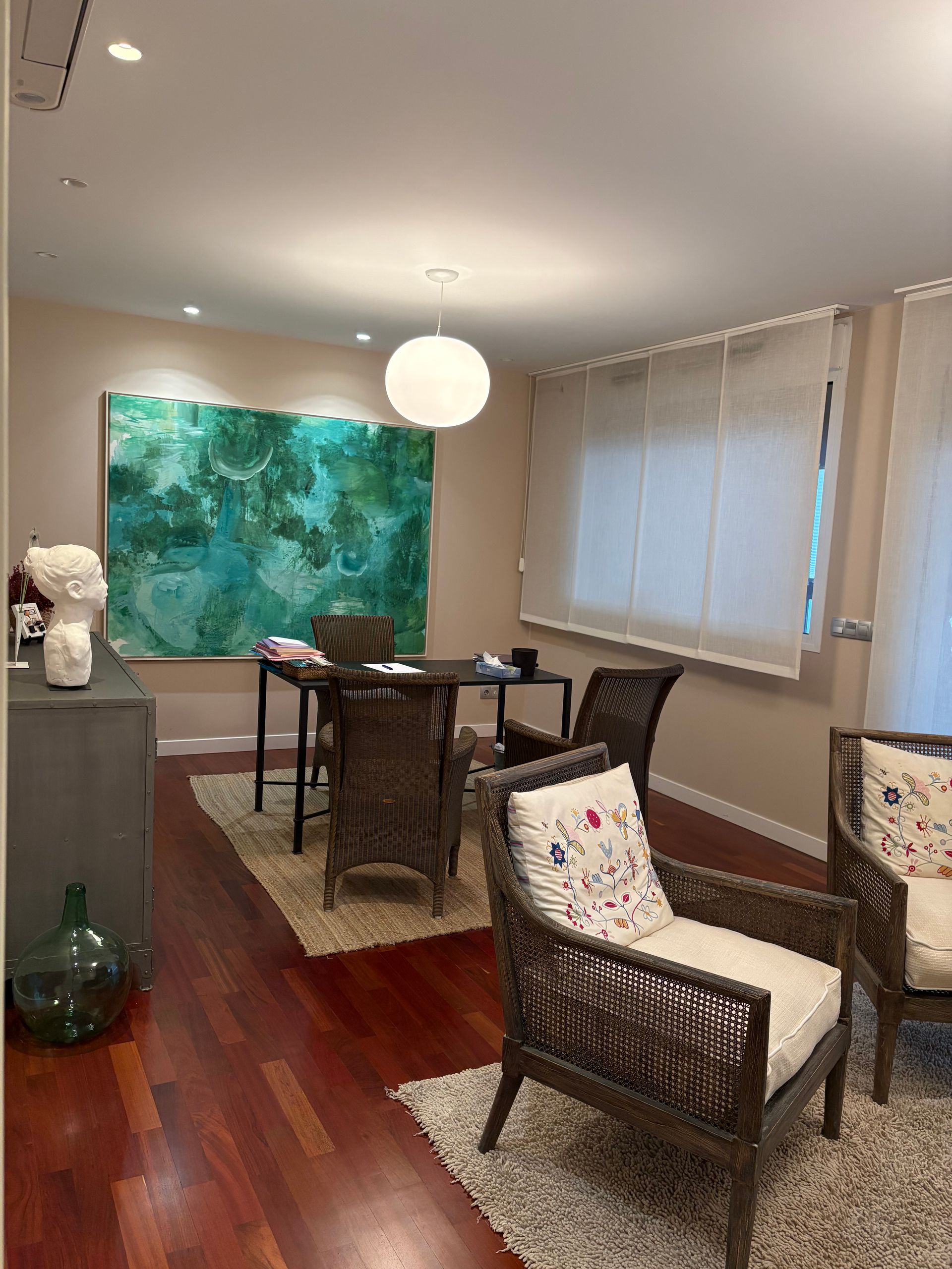 Despacho con un gran cuadro color verde azulado, una mesa con sillas, dos sillas de mimbre y una lámpara colgante redonda.