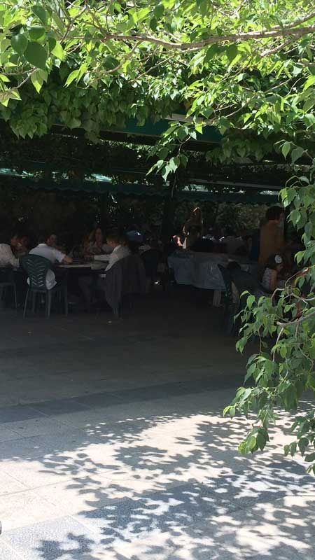 Comedor al aire libre a la sombra de los árboles; la gente se sienta en las mesas.