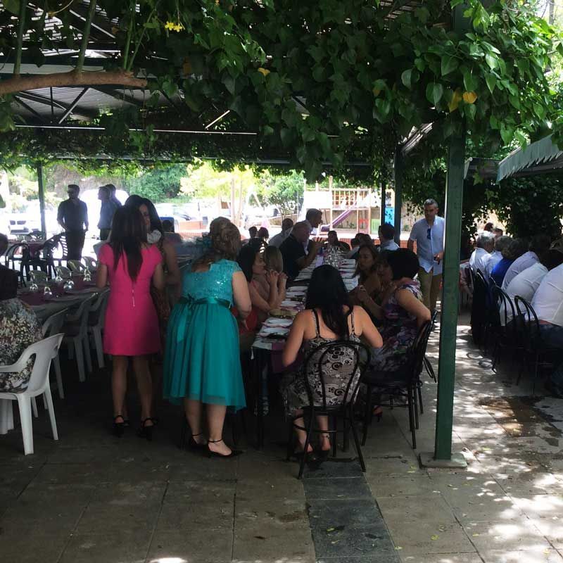 Personas sentadas en mesas al aire libre, bajo un dosel frondoso; algunos permanecen de pie.