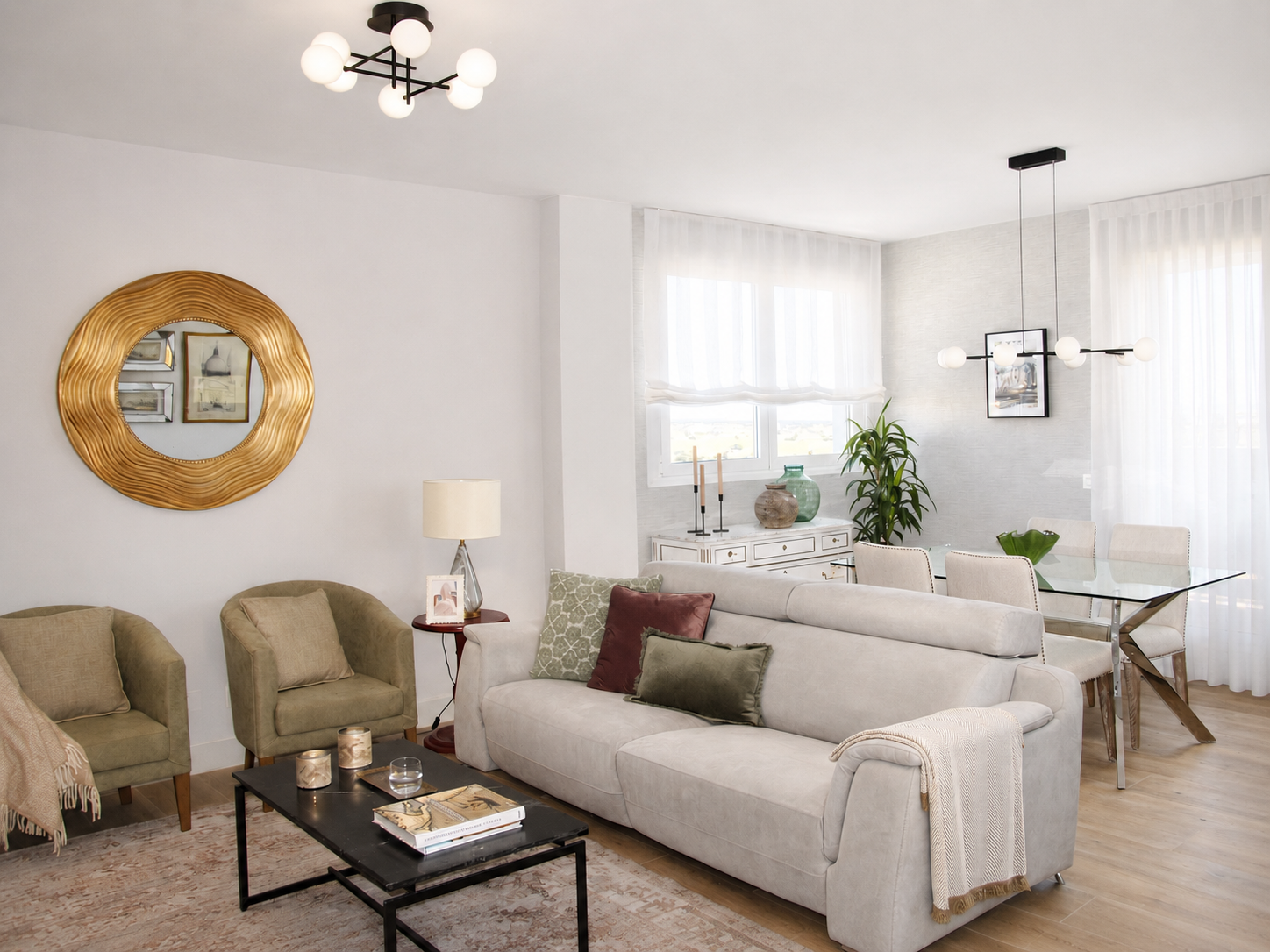 Diseño de salón comedor moderno y luminoso con sofá gris, butacas verdes, mesa de centro con tapa de mármol, espejo redondo dorado y comedor junto a grandes ventanales con cortinas blancas.