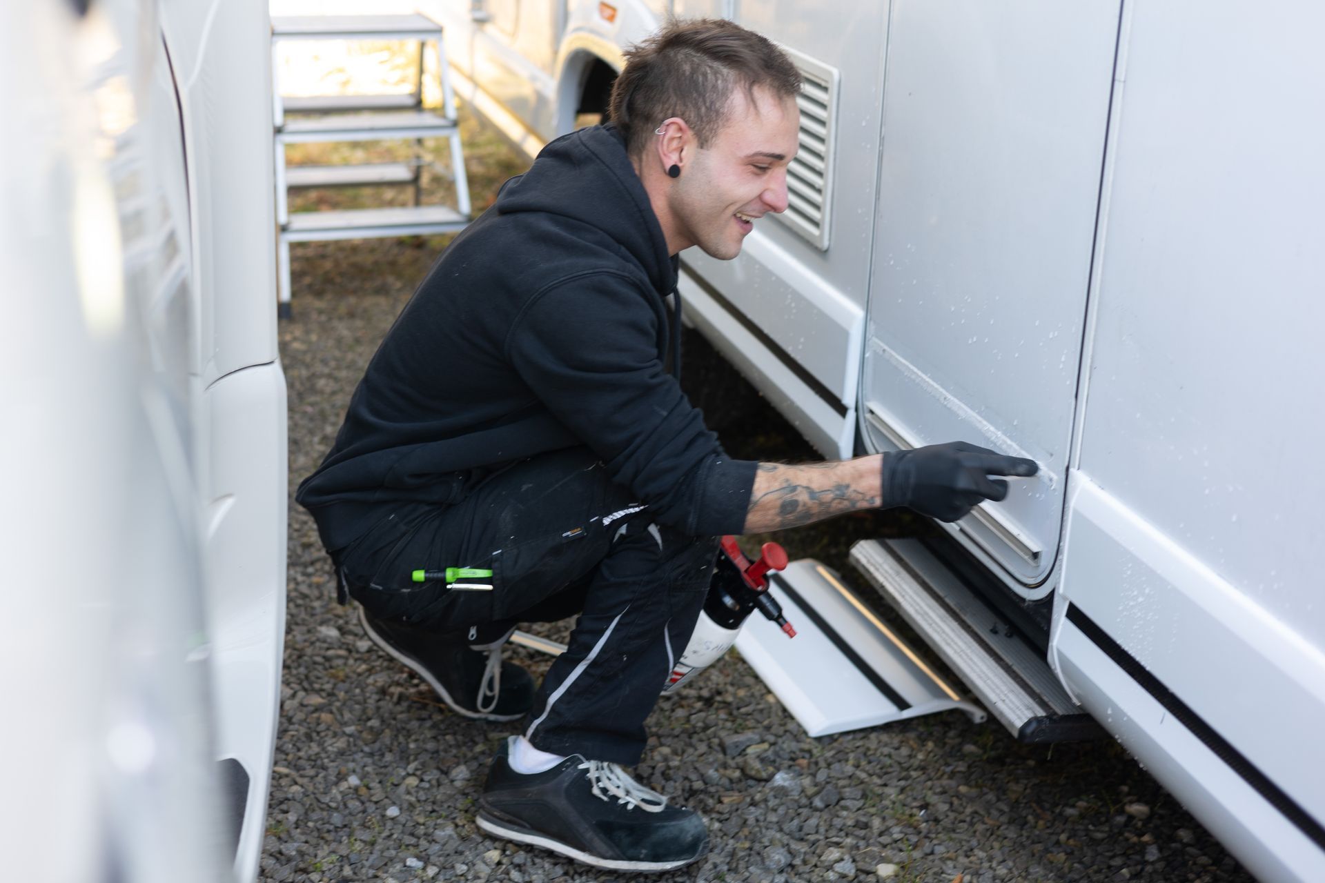 Ein Mann hockt neben einem weißen Wohnmobil und untersucht eine Seitenwand. Er trägt schwarze Arbeitskleidung, schwarze Handschuhe und lächelt.