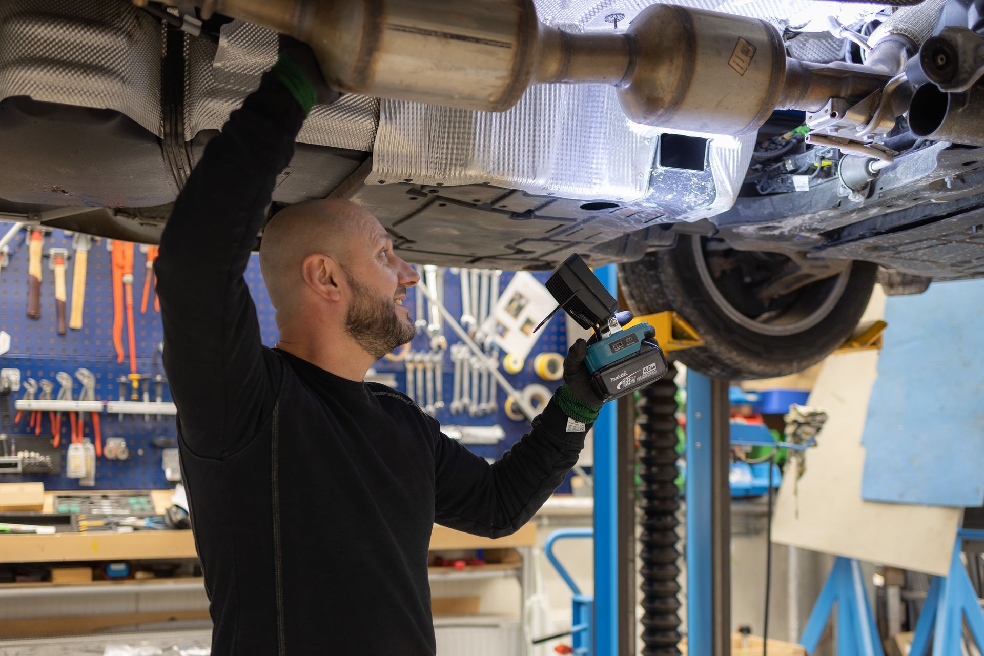 Ein Mechaniker arbeitet in einer Werkstatt unter einem Auto und untersucht mit einer Taschenlampe die Abgasanlage.