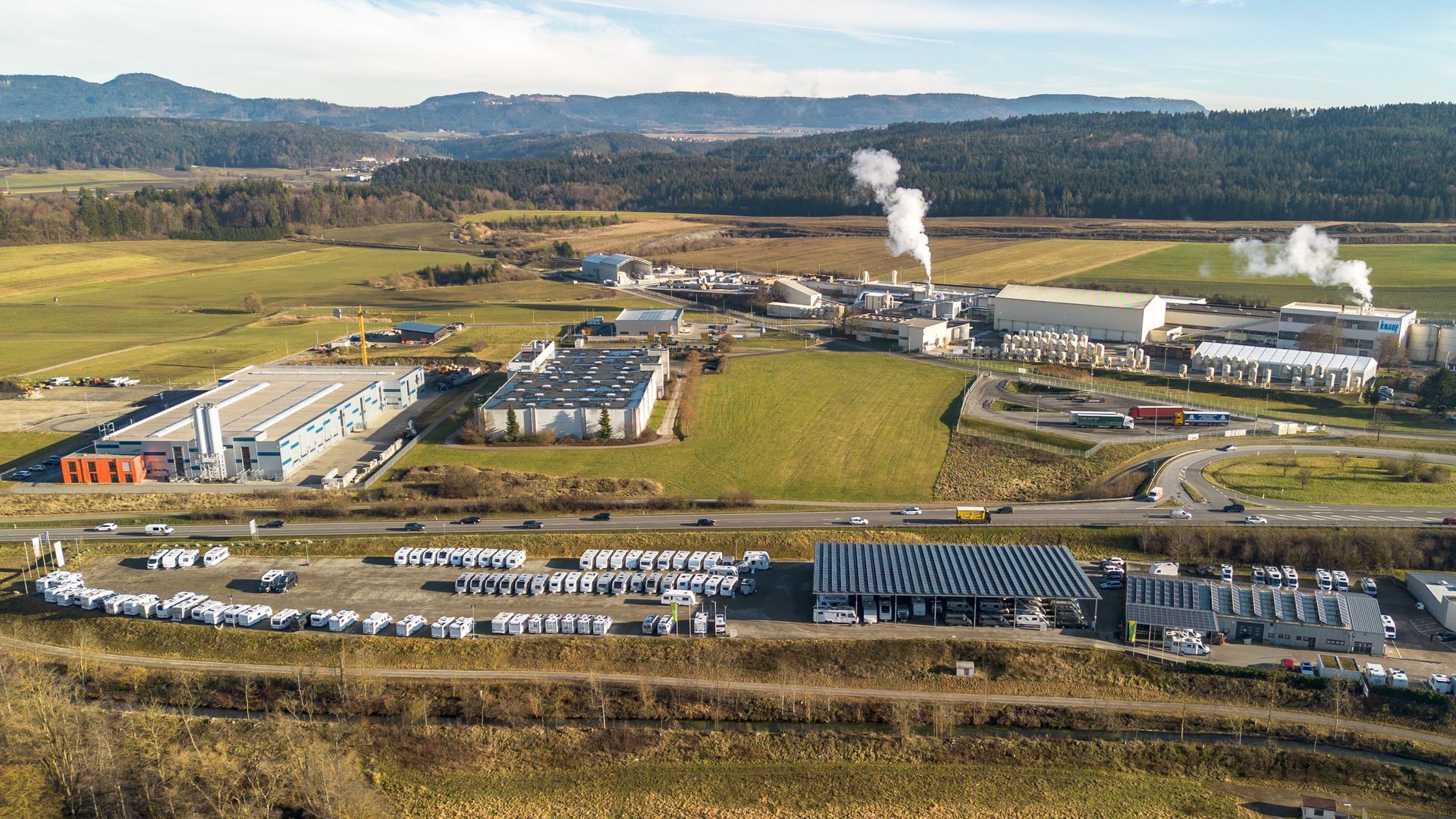 Luftaufnahme eines Industriekomplexes mit Gebäuden, Feldern und einer Straße, aus dem Dampf aufsteigt.