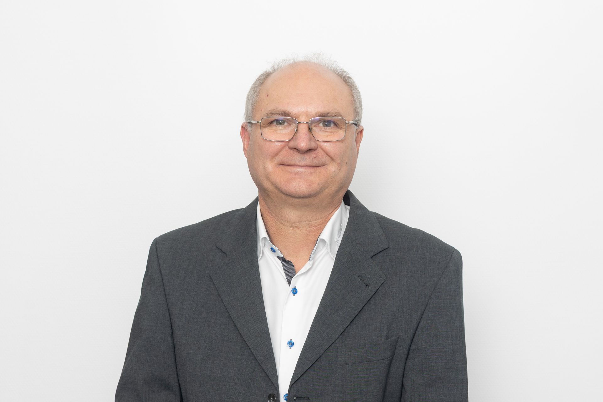 Mann mit Brille und Anzugjacke, lächelnd vor weißem Hintergrund.