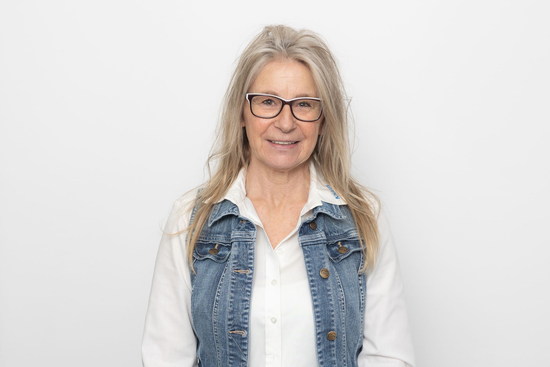 Eine Frau mit Brille, Jeansweste und weißem Hemd lächelt vor einem weißen Hintergrund in die Kamera.