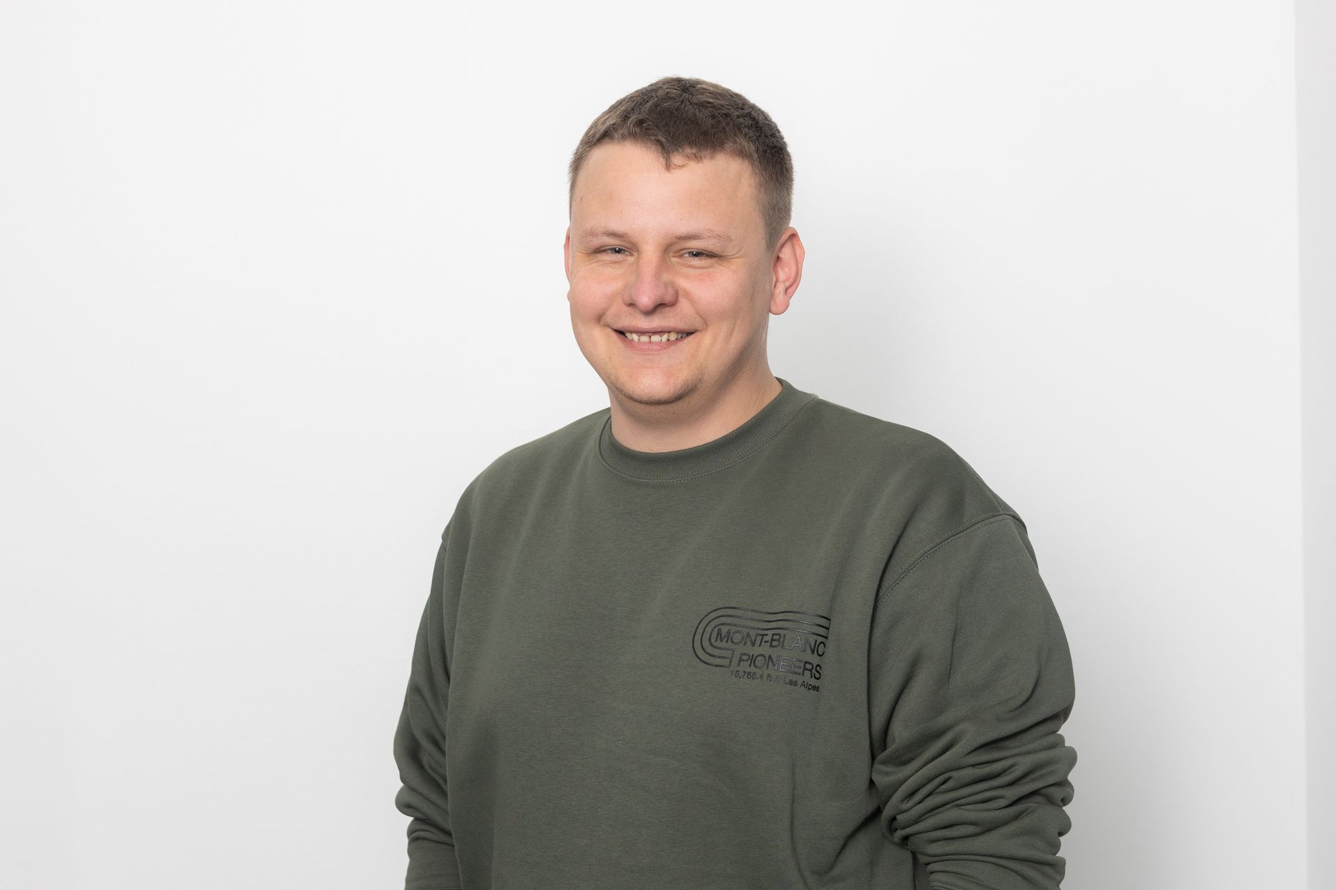 Ein Mann in einem grünen Pullover lächelt vor einer weißen Wand in die Kamera.