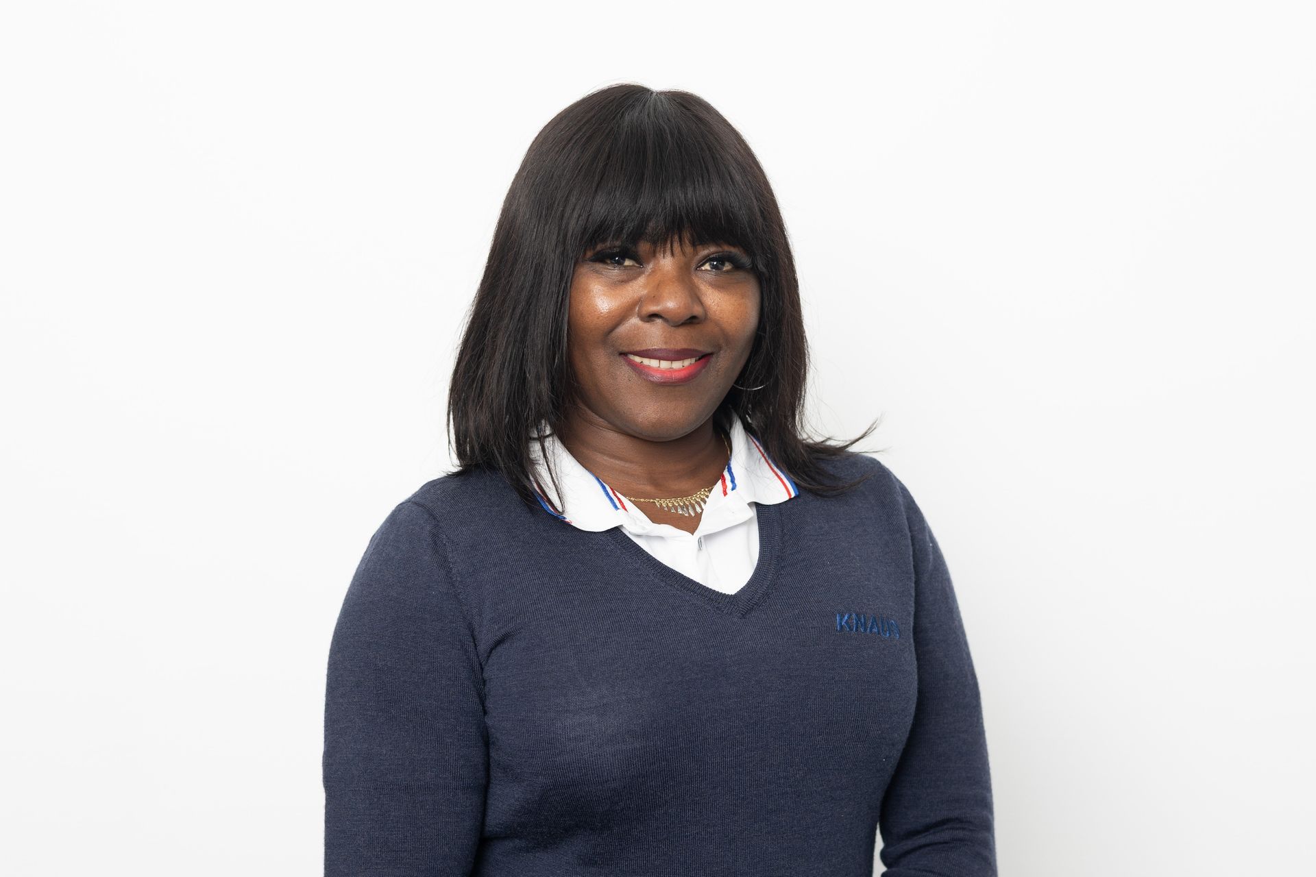 Frau lächelt in einem blauen Pullover und weißem Hemd vor weißem Hintergrund.