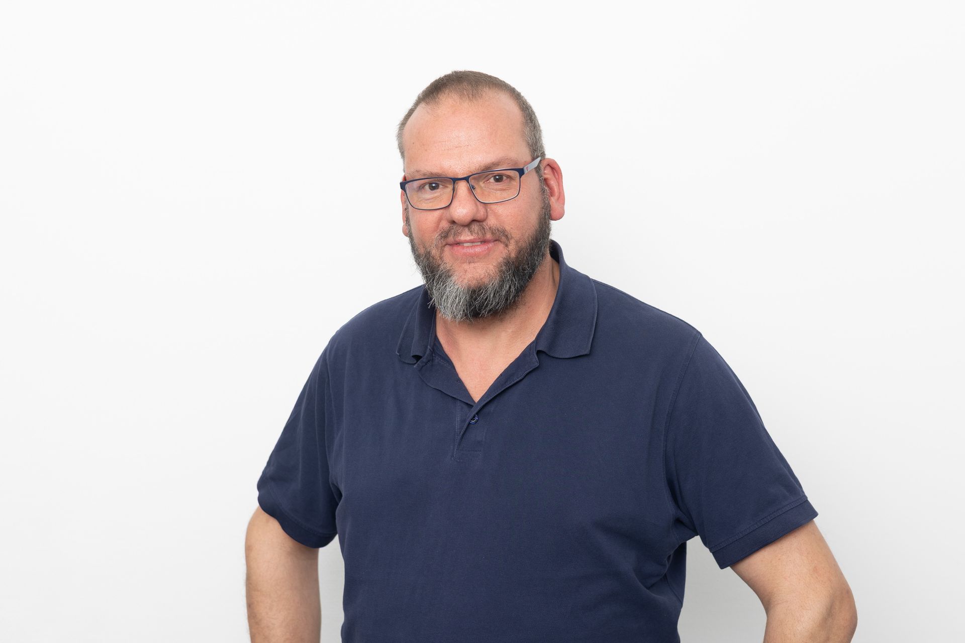 Mann mit Bart, Brille und blauem Poloshirt vor weißem Hintergrund.