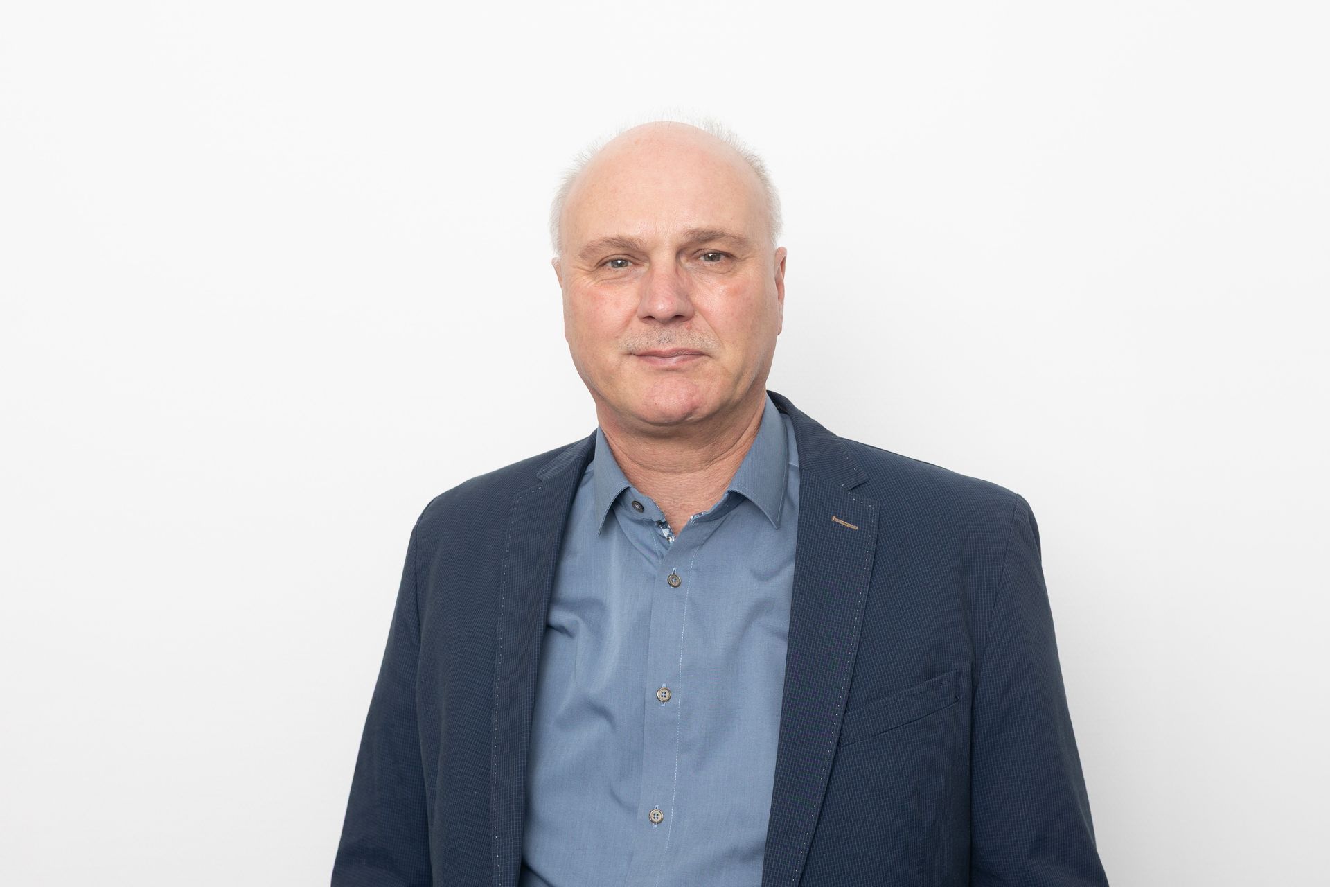 Ein Mann mit kurzen Haaren, der einen blauen Blazer und ein zugeknöpftes Hemd trägt, steht vor einer weißen Wand.
