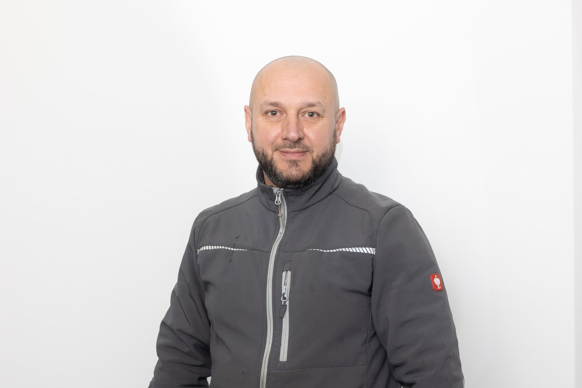 Mann mit kahlgeschorenem Kopf und Bart in einer grauen Jacke, lächelt vor einem weißen Hintergrund.