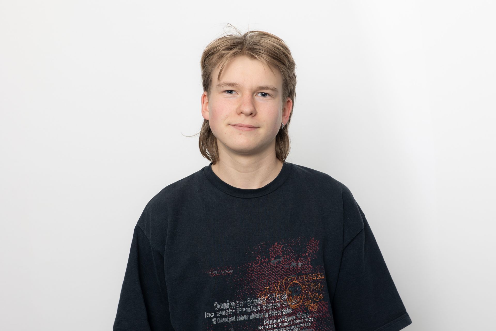 Person mit blonder Vokuhila-Frisur, die ein dunkles T-Shirt trägt, steht vor einem weißen Hintergrund.