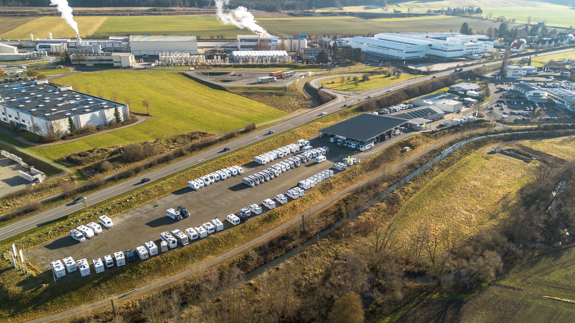 Luftaufnahme eines Fahrzeuglagerplatzes neben einer Autobahn, im Hintergrund Industriegebäude und Felder.