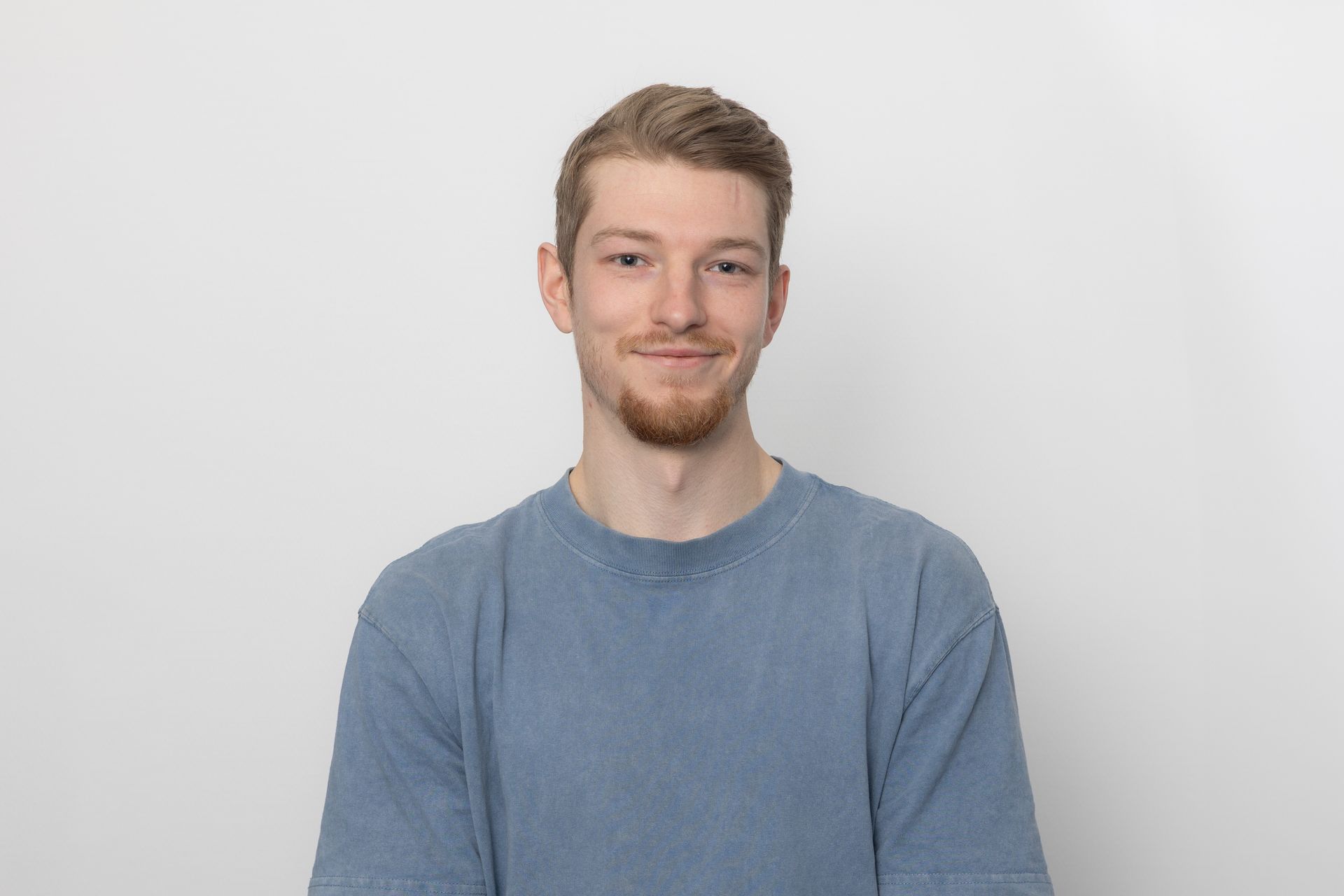 Ein Mann mit hellen Haaren und Bart, der einen blauen Pullover trägt, lächelt vor einem weißen Hintergrund in die Kamera.