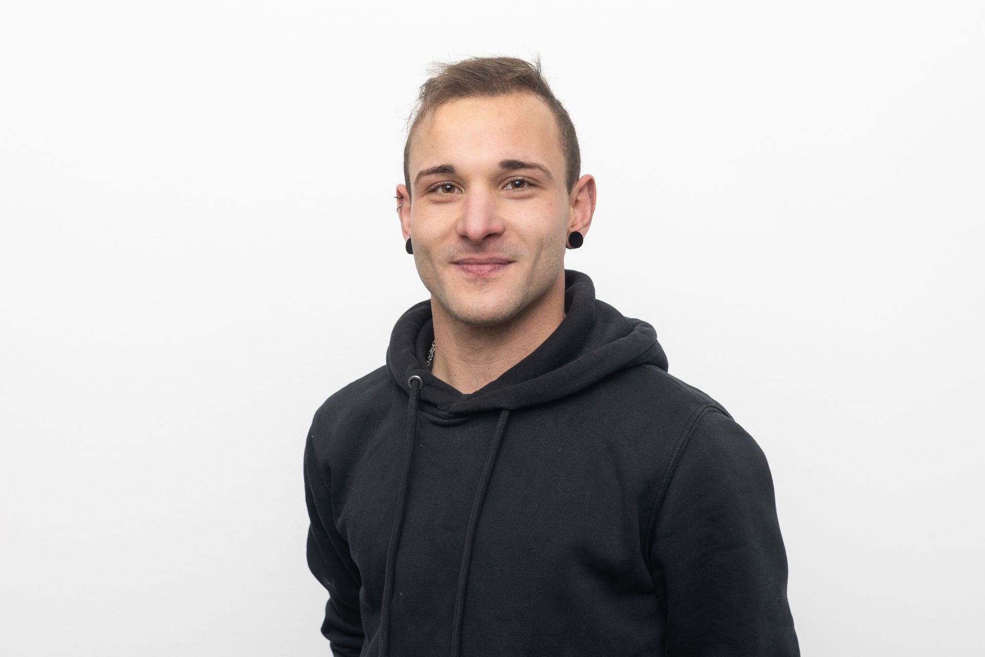Mann mit kurzen braunen Haaren, Ohrlöchern und schwarzem Kapuzenpulli, lächelt vor weißem Hintergrund.