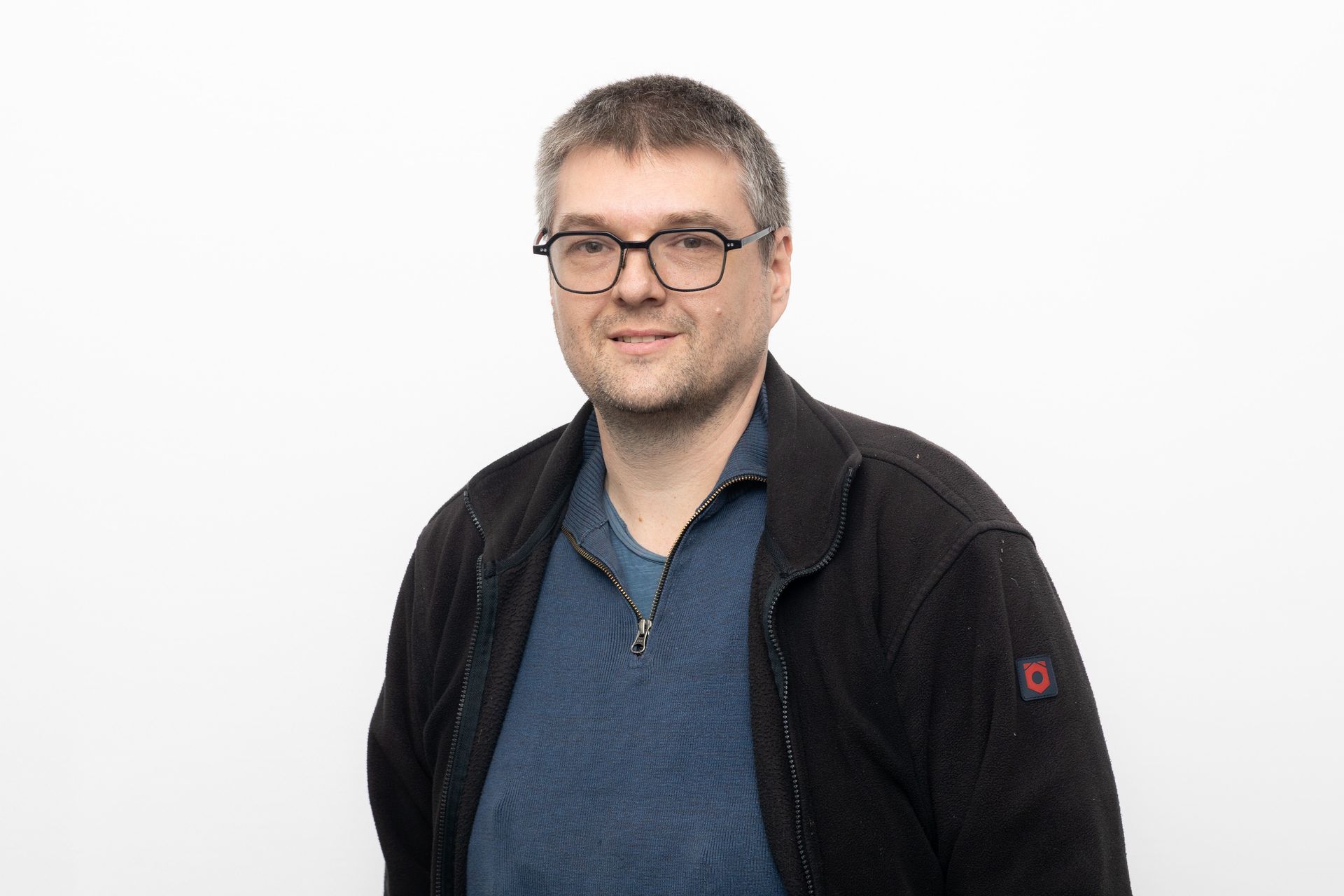Ein Mann mit Brille, blauem Hemd und schwarzer Jacke lächelt vor einem weißen Hintergrund.
