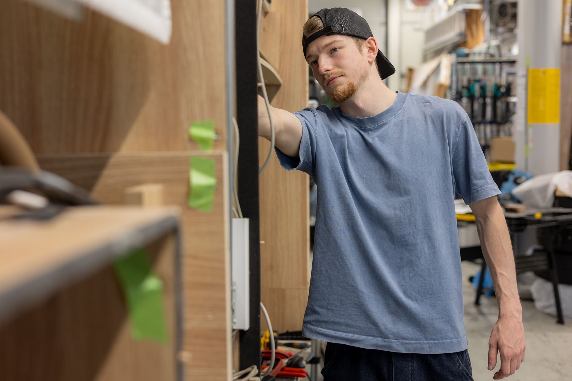 Ein Mann in einer Werkstatt, der an einem Holzschrank die Verkabelung justiert; er trägt eine umgedrehte Baseballkappe und ein blaues Hemd.