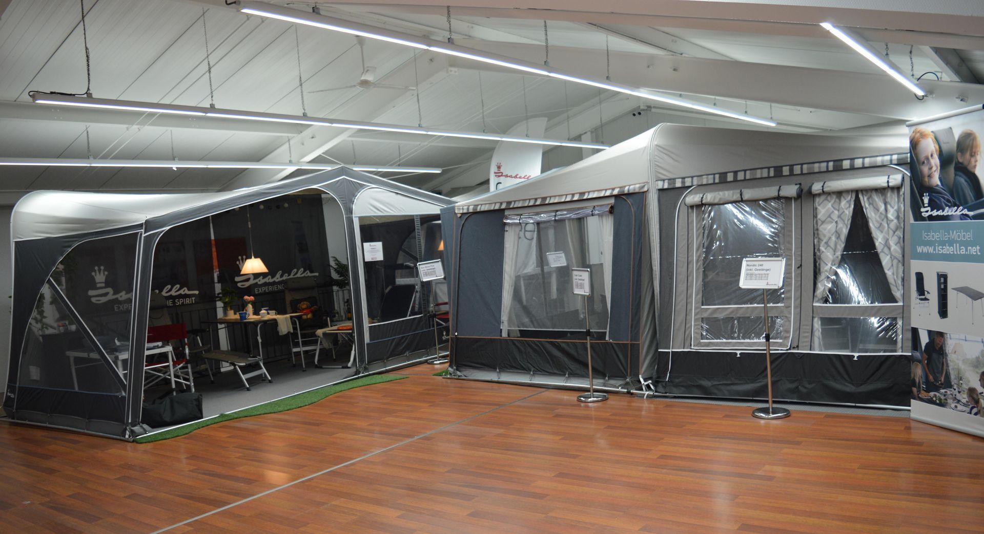 Zwei Campingzelte mit Holzboden sind in einem Innenraum ausgestellt. Eines ist grau mit transparenten Fenstern, das andere schwarz.