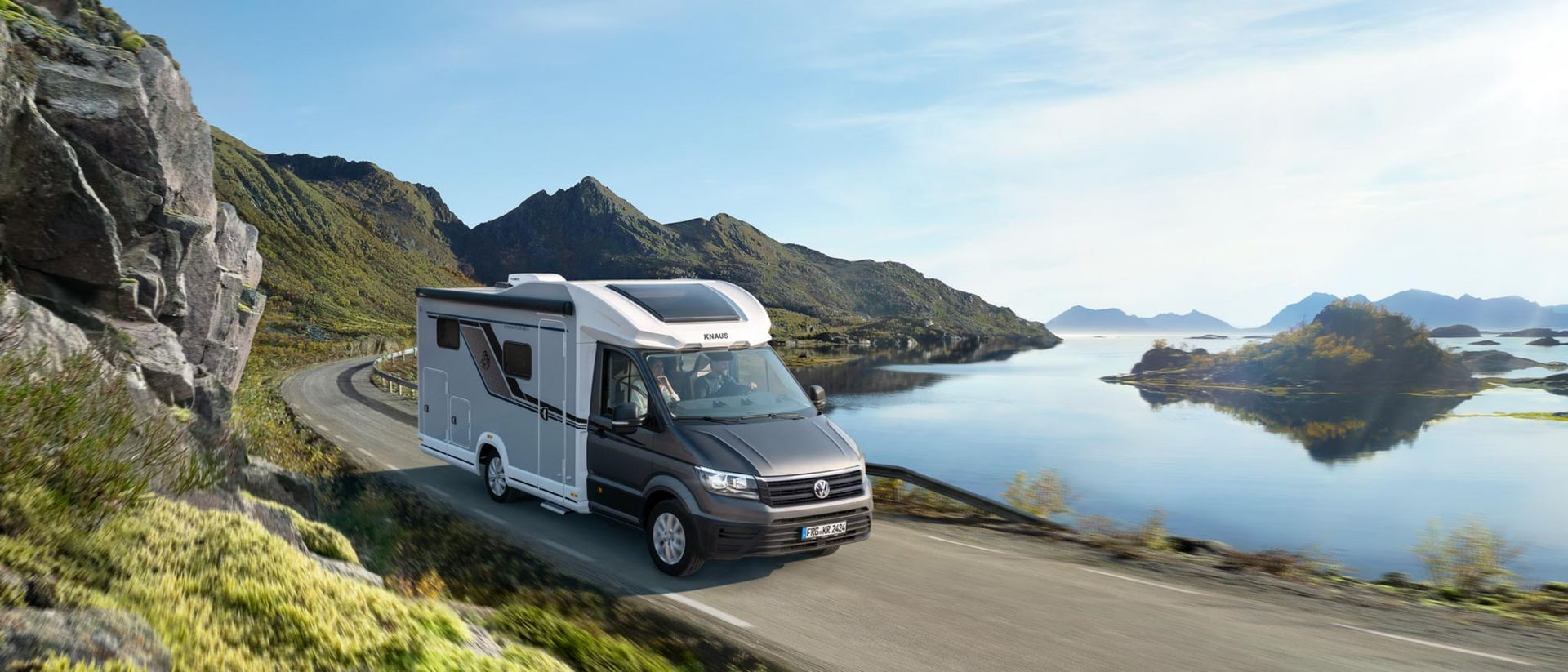Das Wohnmobil fährt auf einer Küstenstraße, im Hintergrund sind Berge und Wasser zu sehen.