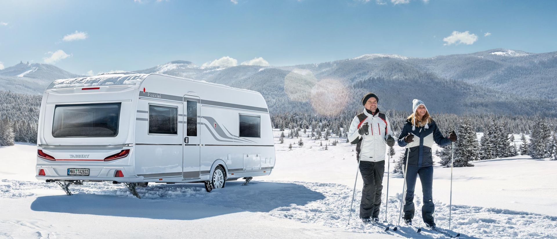 Zwei Personen mit Skiern stehen im Schnee neben einem weißen Wohnmobil vor einer schneebedeckten Bergkulisse.