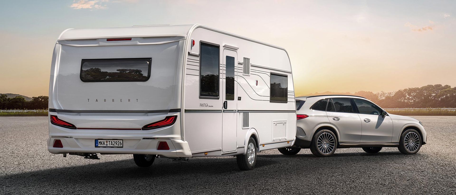 Ein weißer Geländewagen zieht einen weißen Wohnwagen auf einer asphaltierten Straße. Im Hintergrund ist ein Sonnenuntergang über einem Gewässer zu sehen.