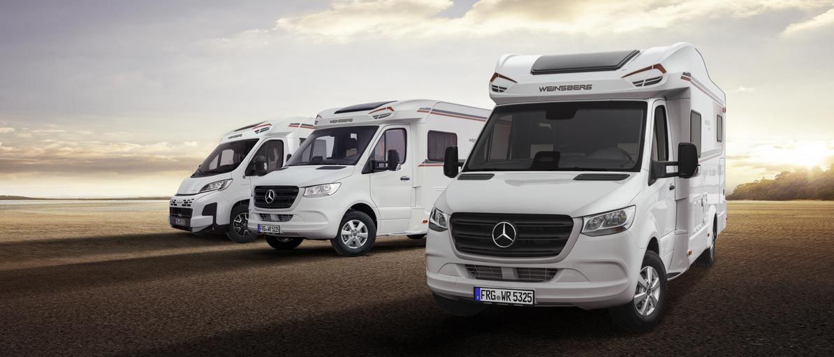 Vier weiße Wohnmobile parkten auf einem gepflasterten Platz vor der Kulisse eines Sonnenuntergangs.