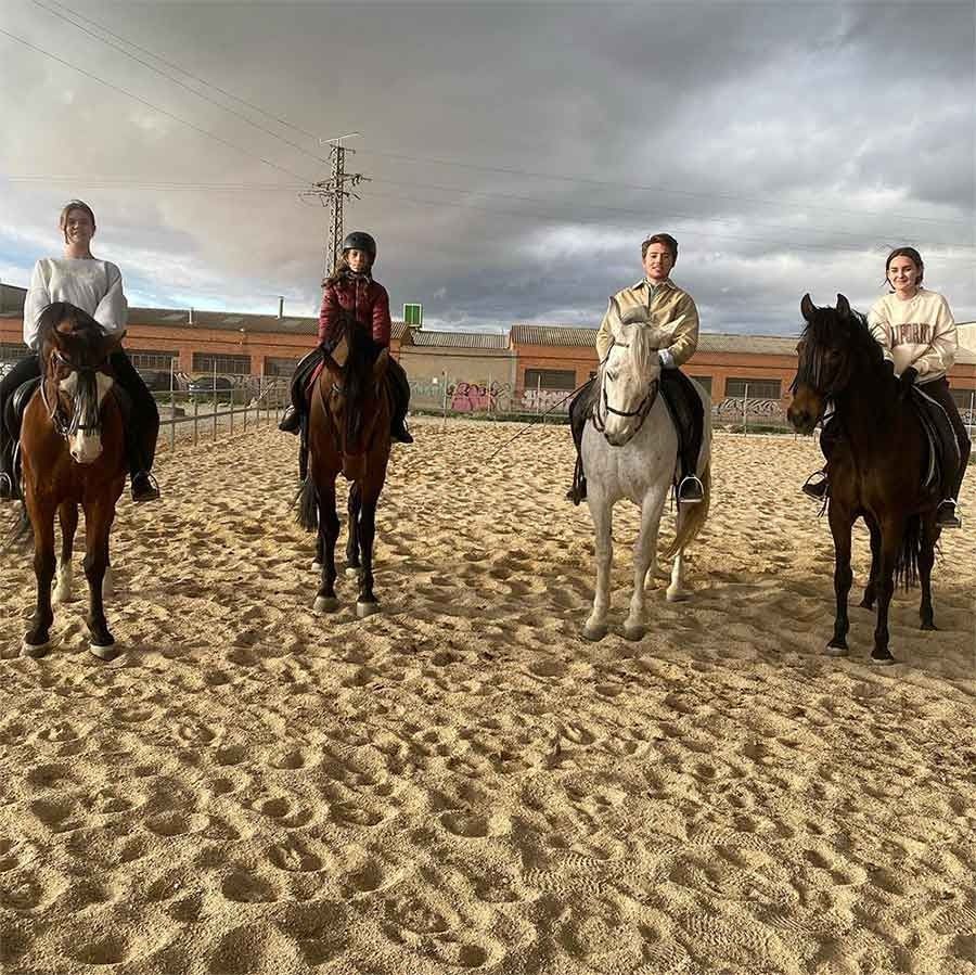 Cuatro personas montando a caballo en una pista de arena bajo un cielo nublado.