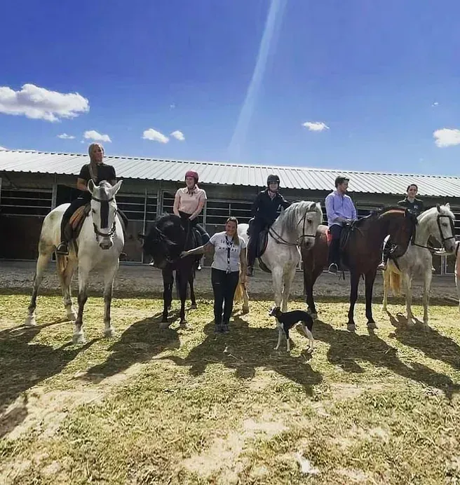 Cinco jinetes a caballo y una persona de pie con perros en un soleado patio de establos al aire libre.