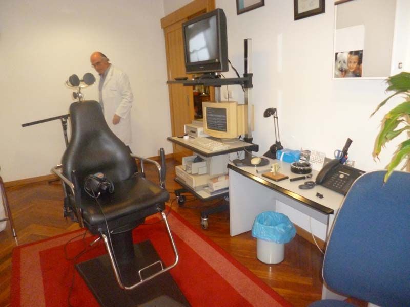 Sala de exploración de una clínica oftalmológica con un médico junto a una silla de exploración negra y equipo médico.