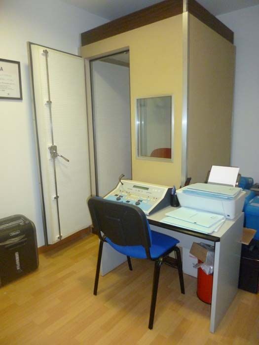 Pequeño cubículo de oficina con escritorio, silla, equipo informático e impresora en una habitación con suelo de madera.
