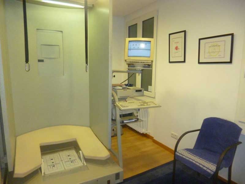 Pequeña sala de consulta con una silla, una camilla de exploración y un monitor médico sobre un carrito.