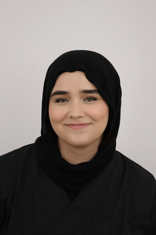 Porträt einer Frau mit schwarzem Kopftuch, die vor einem hellgrauen Hintergrund lächelt