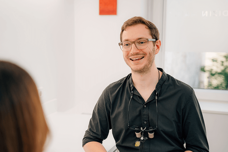 Eine lächelnde Person in schwarzem Hemd und Brille sitzt in einem hellen Büro.