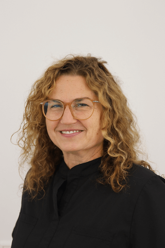Porträt einer lächelnden Person mit lockigem blonden Haar und Brille, die ein schwarzes Oberteil trägt, vor weißem Hintergrund