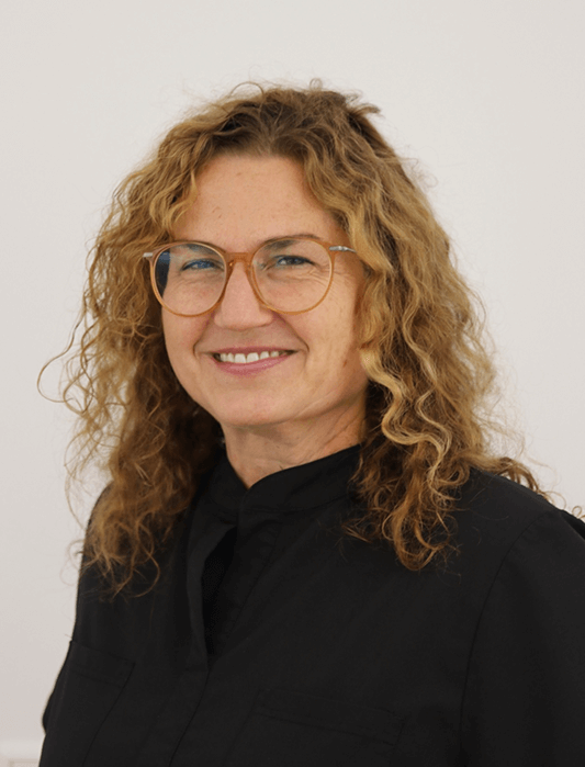 Porträt einer lächelnden Person mit lockigem blonden Haar und Brille, die ein schwarzes Oberteil trägt, vor hellem Hintergrund