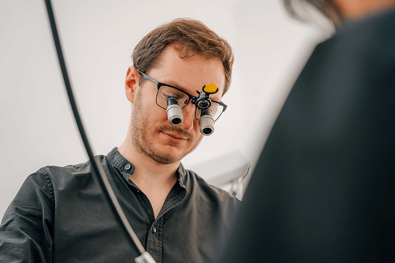 Ein Techniker mit Vergrößerungsbrille justiert Geräte in einer hellen Klinikumgebung.